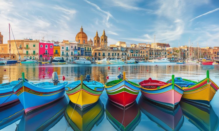 Traditional colorful luzzu boats are floating on the water with the skyline of marsaxlokk in the background